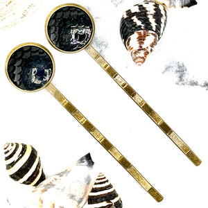 All Up In The Hair | Online Accessory Boutique Located in Mooresville, NC | Two Black Mermaid Bobby Pins laying on a white marble background. There are multiple shells surrounding the bobby pins.