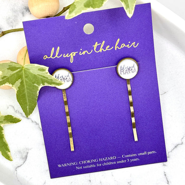 All Up In The Hair | Online Accessory Boutique Located in Mooresville, NC | Two Blessed Mama Bobby Pins on an indigo colored, All Up In The Hair branded packaging card. The card is laying on a white marble background. There is a wood bead garland and ivy leaves next to the card.
