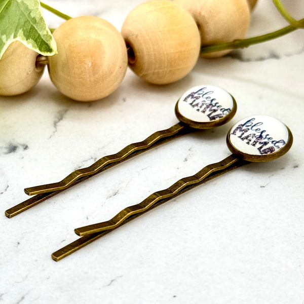 All Up In The Hair | Online Accessory Boutique Located in Mooresville, NC | Side view of two Blessed Mama Bobby Pins laying on a white marble background. There is a wood bead garland and ivy leaves next to the bobby pins.