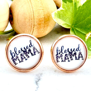 All Up In The Hair | Online Accessory Boutique Located in Mooresville, NC | Two round white earrings with the words "blessed mama" written on them. The earrings are laying on a white marble background. There is a wood bead garland and ivy leaves behind the earrings.