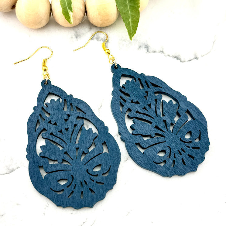 All Up In The Hair | Online Accessory Boutique Located in Mooresville, NC | Two Blue Floral Wood Dangle Earrings on a white marble background. There is a wood bead garland and ivy leaves behind the earrings.