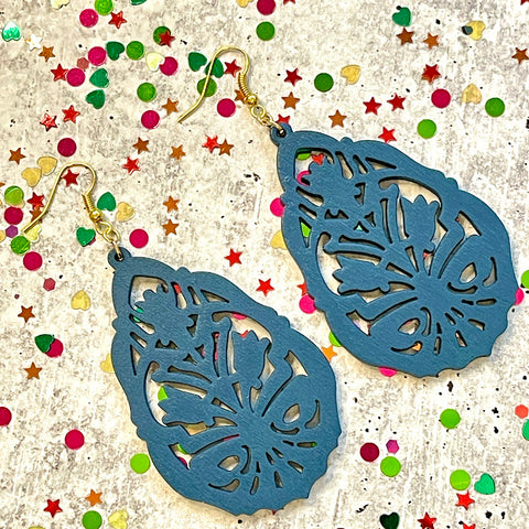 All Up In The Hair | Online Accessory Boutique Located in Mooresville, NC | Two laser cut wood earrings, painted blue, are laying on a gray background, surrounded by colorful glitter.