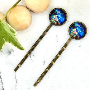 All Up In The Hair | Online Accessory Boutique Located in Mooresville, NC | Two Blue Galaxy Bobby Pins on a white marble background. There is a wood bead garland and ivy leaves next to the bobby pins.