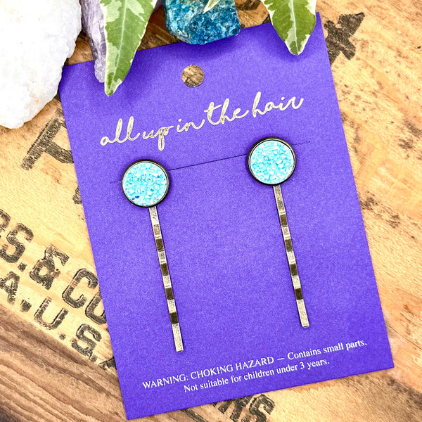 All Up In The Hair | Online Accessory Boutique Located in Mooresville, NC | Two light blue druzy bobby pins on an indigo colored, All Up In The Hair branded packaging card. The card is laying on a wood background with black lettering. There are crystals and ivy leaves at the top of the image.