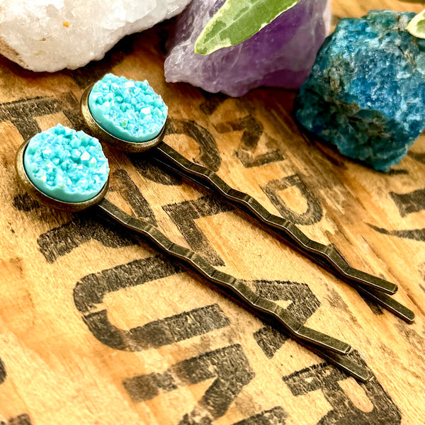 All Up In The Hair | Online Accessory Boutique Located in Mooresville, NC | Two Blue Lace Agate Druzy Bobby Pins laying diagonally on a wood background with black lettering. There are crystals and ivy leaves at the top of the image.