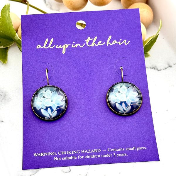 All Up In The Hair | Online Accessory Boutique Located in Mooresville, NC | Two Blue Lily Dangle Earrings on an indigo colored, All Up In The Hair branded packaging card. The card is laying on a white marble background.