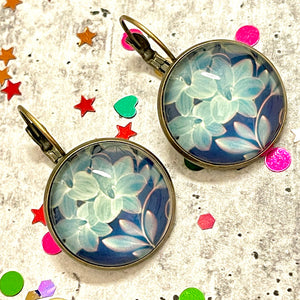 All Up In The Hair | Online Accessory Boutique Located in Mooresville, NC | Two blue lily dangle earrings laying on a gray background, surrounded by colorful glitter.