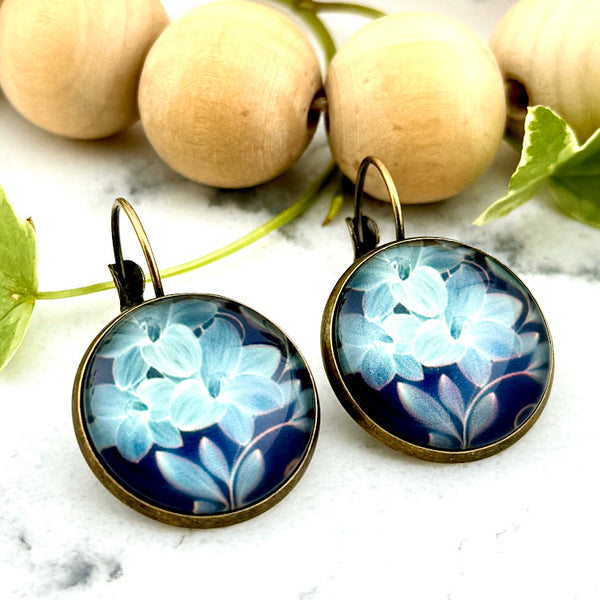 All Up In The Hair | Online Accessory Boutique Located in Mooresville, NC | Two Blue Lily Dangle Earrings on a white marble background. Behind the earrings is a wood bead garland and ivy leaves.