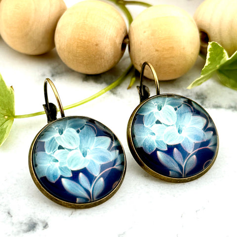 All Up In The Hair | Online Accessory Boutique Located in Mooresville, NC | Two Blue Lily Dangle Earrings on a white marble background. Behind the earrings is a wood bead garland and ivy leaves.