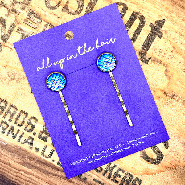 All Up In The Hair | Online Accessory Boutique Located in Mooresville, NC | Two Blue Mermaid Bobby Pins on an indigo colored, All Up In The Hair branded packaging card. The card is laying on a wood background with black lettering.