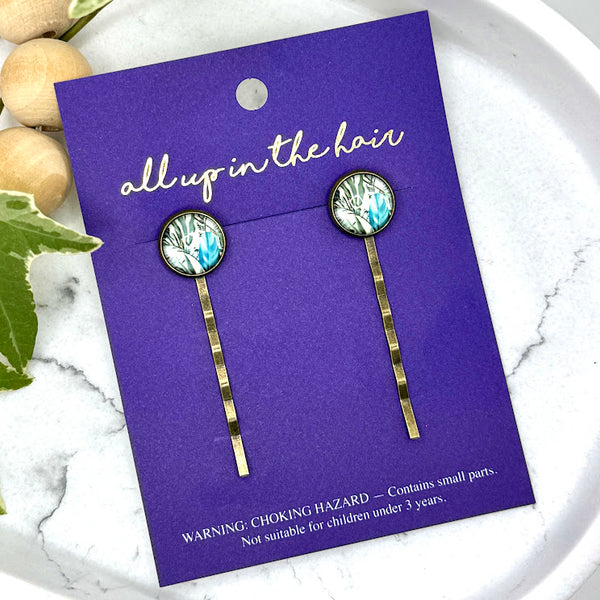 All Up In The Hair | Online Accessory Boutique Located in Mooresville, NC | Two Blue Paradise Bobby Pins on an indigo colored, All Up In The Hair branded packaging card. The card is laying on a white marble background next to a wood bead garland and ivy leaves.