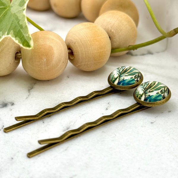 All Up In The Hair | Online Accessory Boutique Located in Mooresville, NC | Side view of two Blue Paradise Bobby Pins on a white marble background. There is a wood bead garland and ivy leaves next to the bobby pins.