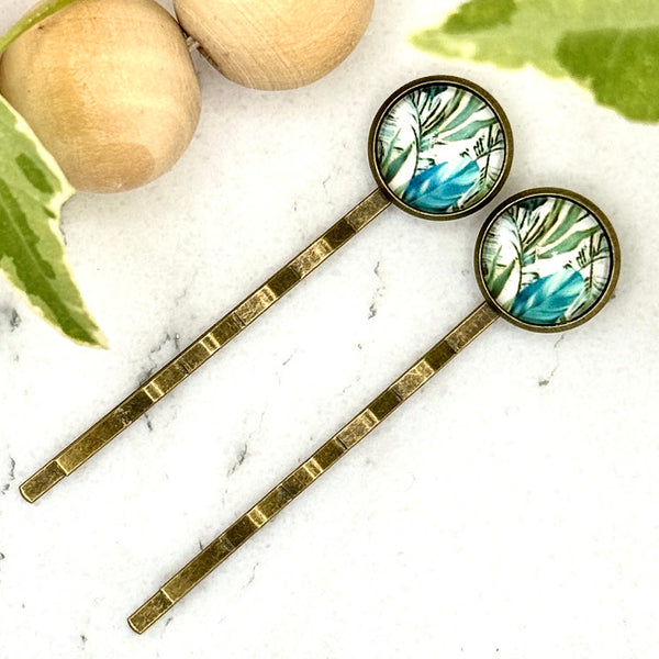 All Up In The Hair | Online Accessory Boutique Located in Mooresville, NC | Two bobby pins with tropical leaves and a blue flower on a white marble background. There is a wood bead garland and ivy leaves next to the bobby pins.