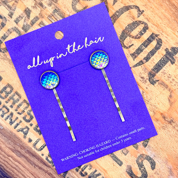 All Up In The Hair | Online Accessory Boutique Located in Mooresville, NC | Two Bright Purple Mermaid Bobby Pins on an indigo colored, All Up In The Hair packaging card. The card is laying on a wood background with black lettering.