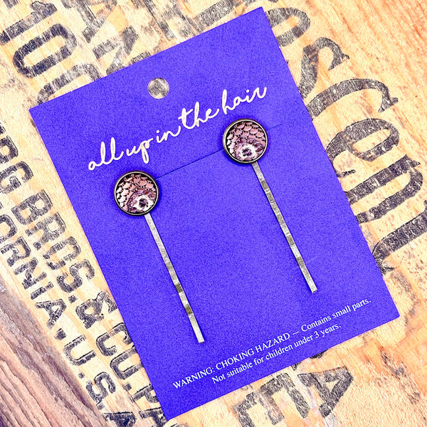 All Up In The Hair | Online Accessory Boutique Located in Mooresville, NC | Two Bronze Mermaid Bobby Pins on an indigo-colored, All Up In The Hair branded packaging card. The card is laying on a wood background with black lettering.