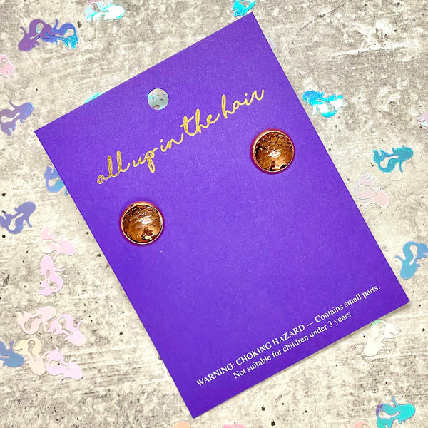 All Up In The Hair | Online Accessory Boutique Located in Mooresville, NC | An indigo colored, All Up In The Hair branded packaging card laying on a gray background, surrounded by colorful glitter. On the card is two bronze mermaid earrings.