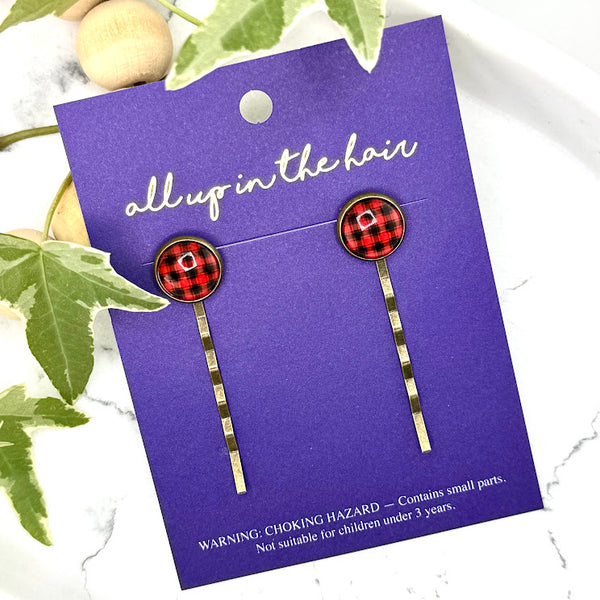 All Up In The Hair | Online Accessory Boutique Located in Mooresville, NC | Two Buffalo Plaid Bobby Pins on an indigo colored, All Up In The Hair branded packaging card. The card is laying on a white marble background next to a wood bead garland and ivy leaves.