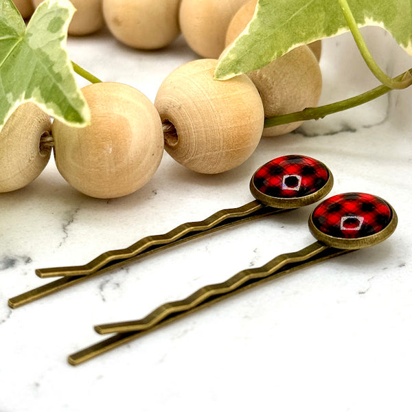 All Up In The Hair | Online Accessory Boutique Located in Mooresville, NC | Side view of two Buffalo Plaid Bobby Pins laying on a white marble background next to a wood bead garland and ivy leaves.