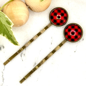 All Up In The Hair | Online Accessory Boutique Located in Mooresville, NC | Two red Buffalo Plaid Bobby Pins laying on a white marble background next to a wood bead garland and ivy leaves.