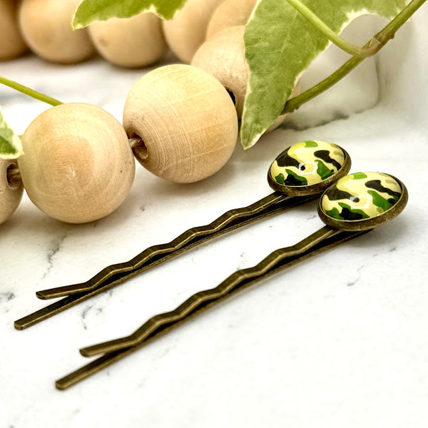 All Up In The Hair | Online Accessory Boutique Located in Mooresville, NC | Side view of two Camo Bobby Pins laying on a white marble background next to a wood bead garland and ivy leaves.