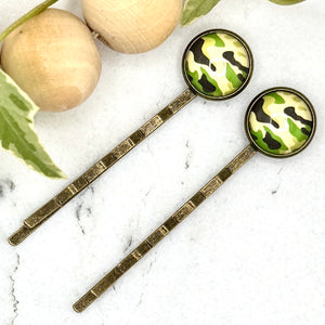 All Up In The Hair | Online Accessory Boutique Located in Mooresville, NC | Two Camo Bobby Pins laying on a white marble background next to a wood bead garland and ivy leaves.