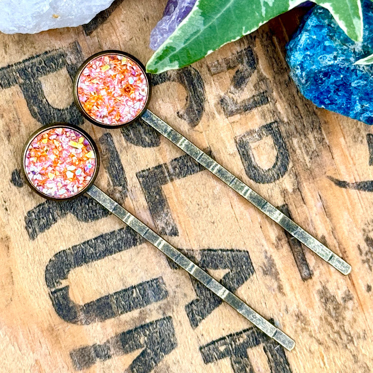 All Up In The Hair | Online Accessory Boutique Located in Mooresville, NC | Two Carnelian druzy bobby pins laying diagonally on a grey background, surrounded by colorful glitter.