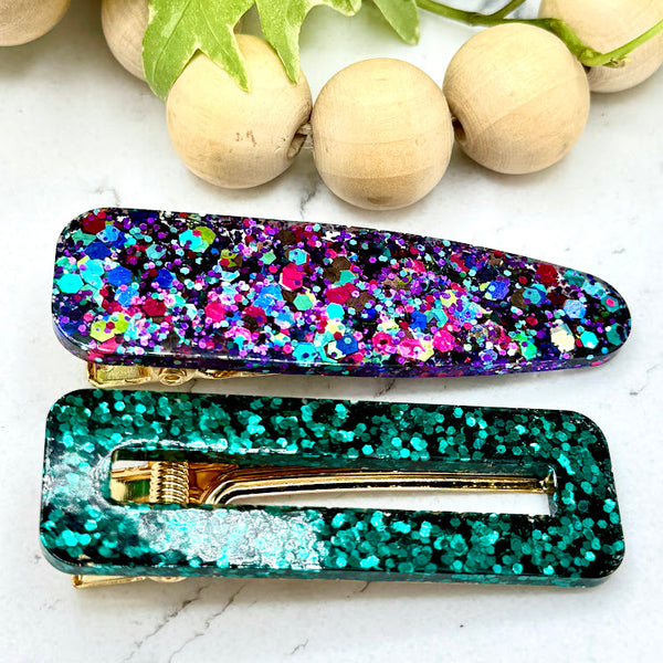 All Up In The Hair | Online Accessory Boutique Located in Mooresville, NC | One emerald green rectangular barrette and one "teardrop" multicolored glitter barrette laying on a grey background surrounded by colorful glitter