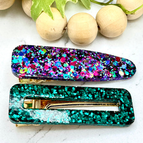 All Up In The Hair | Online Accessory Boutique Located in Mooresville, NC | One teardrop shaped, multicolor glitter barrette and one hollow rectangle, emerald green glitter barrette laying on a white marble background. There is a wood bead garland and ivy leaves at the top of the image.