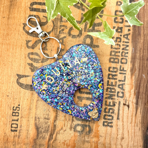 All Up In The Hair | Online Accessory Boutique Located in Mooresville, NC | Celestial Planchette Keychain on a wood background with black lettering. There are ivy leaves next to the keychain.