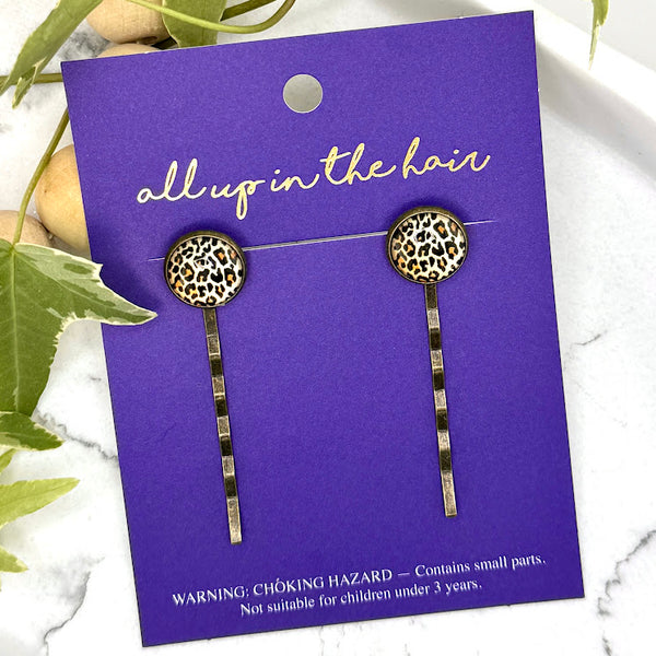 All Up In The Hair | Online Accessory Boutique Located in Mooresville, NC | Two Cheetah Bobby Pins on an indigo colored, All Up In The Hair branded packaging card. The card is laying on a white marble background next to a wood bead garland and ivy leaves.