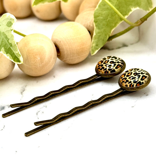 All Up In The Hair | Online Accessory Boutique Located in Mooresville, NC | Side view of two Cheetah Bobby Pins laying on a white marble background next to a wood bead garland and ivy leaves.