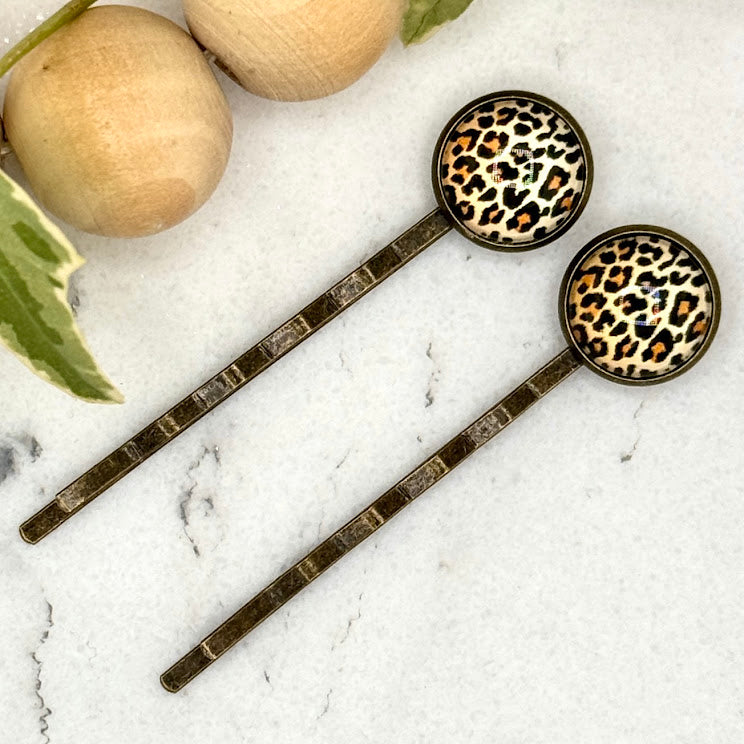 All Up In The Hair | Online Accessory Boutique Located in Mooresville, NC | Two Cheetah Print Bobby Pins laying on a white marble background next to a wood bead garland and ivy leaves.