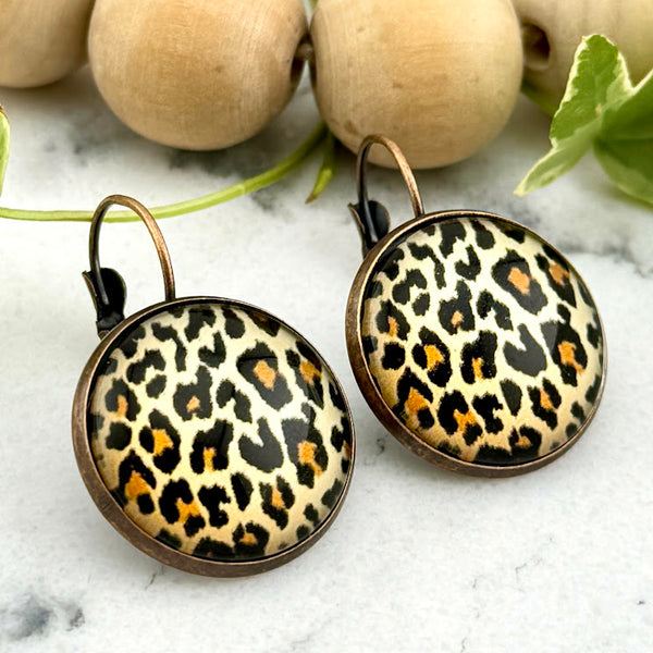 All Up In The Hair | Online Accessory Boutique Located in Mooresville, NC | Two Cheetah Dangle Earrings on a white marble background. Behind the earrings is a wood bead garland and ivy leaves.