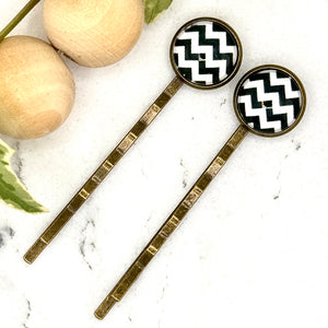 All Up In The Hair | Online Accessory Boutique Located in Mooresville, NC | Two white and black Chevron Bobby Pins laying on a white marble background next to a wood bead garland and ivy leaves.