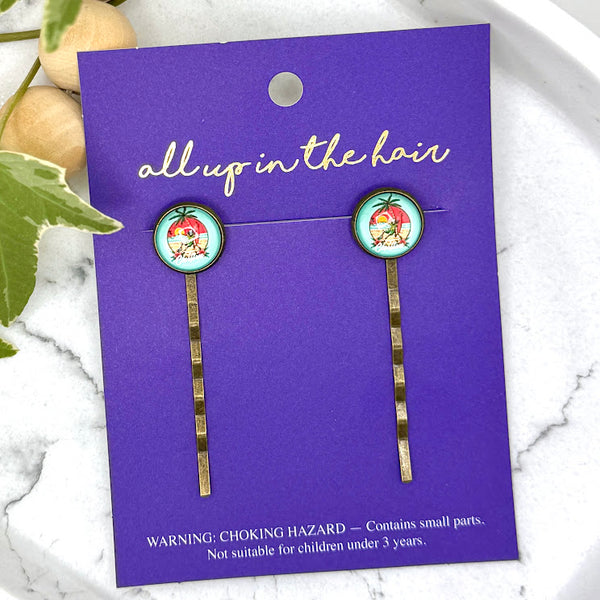 All Up In The Hair | Online Accessory Boutique Located in Mooresville, NC | Two Chillin Skeleton Bobby Pins on an indigo colored, All Up In The Hair branded packaging card. The card is laying on a white marble background next to a wood bead garland and ivy leaves.