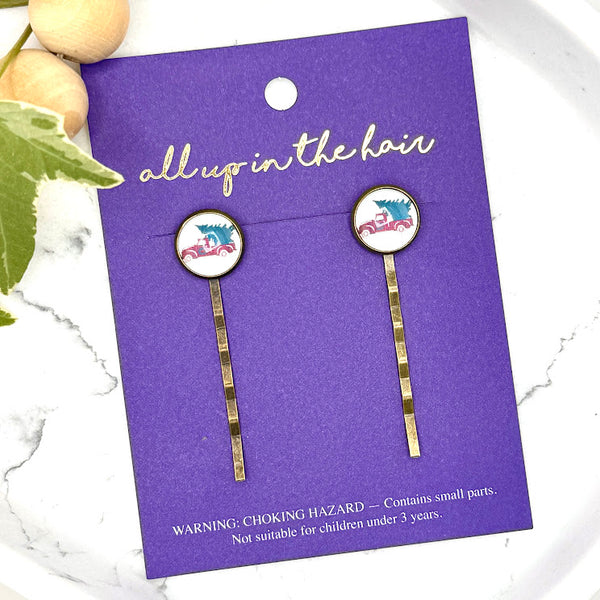 All Up In The Hair | Online Accessory Boutique Located in Mooresville, NC | Two Christmas Tree Truck Bobby Pins on an indigo colored, All Up In The Hair branded packaging card. The card is laying on a white marble background, next to a wood bead garland and ivy leaves.