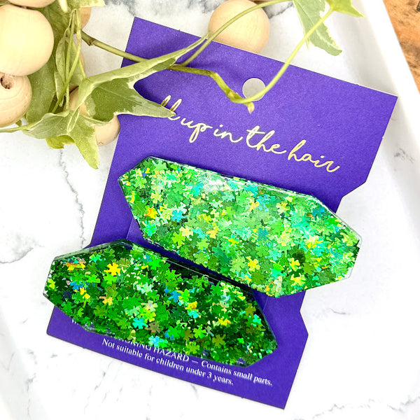 All Up In The Hair | Online Accessory Boutique Located in Mooresville, NC | Our Clover Patch Barrette Set on an indigo colored, All Up In The Hair branded packaging card. The card is laying on a white marble background. At the top of the image is a wood bead garland and ivy leaves.