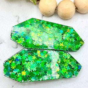 All Up In The Hair | Online Accessory Boutique Located in Mooresville, NC | A green geometric barrette with four leaf clover glitter and a green and gold glitter barrette are laying on a gray background, surrounded by colorful glitter.