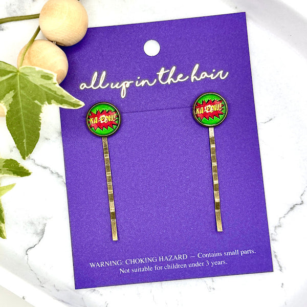 All Up In The Hair | Online Accessory Boutique Located in Mooresville, NC | Two Comic Ka-pow Bobby pins on an All Up In The Hair branded packaging card. The card is laying on a gray background, surrounded by colorful glitter.