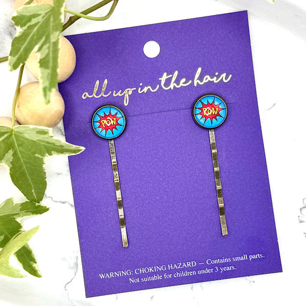All Up In The Hair | Online Accessory Boutique Located in Mooresville, NC | Two Comic Pow Bobby Pins on an indigo colored, All Up In The Hair branded packaging card. The card is laying on a white marble background, next to a wood bead garland and ivy leaves.