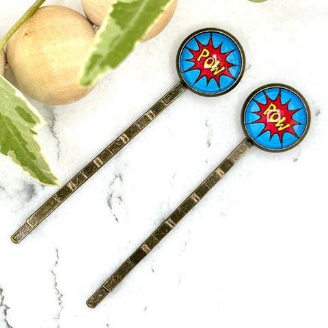 All Up In The Hair | Online Accessory Boutique Located in Mooresville, NC | Two Comic Pow Bobby Pins on a white marble background next to a wood bead garland and ivy leaves.