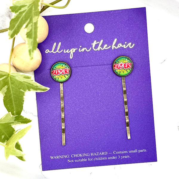 All Up In The Hair | Online Accessory Boutique Located in Mooresville, NC | Two Super Bobby Pins on an All Up In The Hair branded packaging card. The card is laying on a gray background, surrounded by colorful glitter.