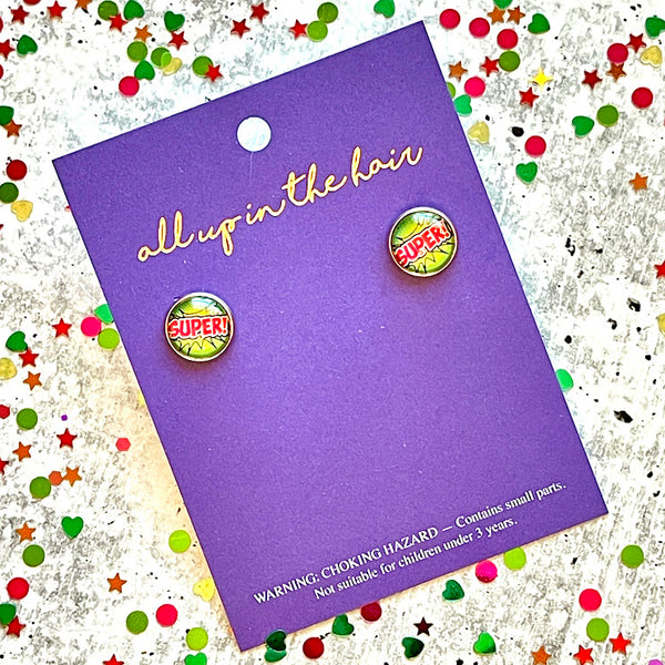 All Up In The Hair | Online Accessory Boutique Located in Mooresville, NC | Two Comic Super earrings on an All Up In The Hair branded packaging card. The card is laying on a gray background, surrounded by colorful glitter.