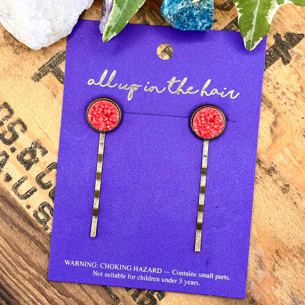 All Up In The Hair | Online Accessory Boutique Located in Mooresville, NC | An All Up In The Hair branded packaging card on a grey background, surrounded by colorful glitter. On the card is two coral druzy bobby pins.