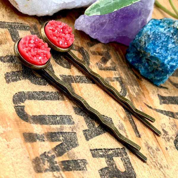 All Up In The Hair | Online Accessory Boutique Located in Mooresville, NC | Two Coral Druzy Bobby Pins laying on top of wood background with black lettering. There are crystals and ivy leaves at the top of the image.