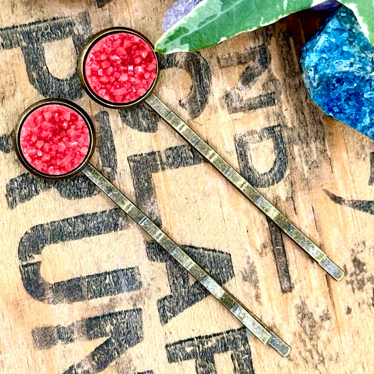 All Up In The Hair | Online Accessory Boutique Located in Mooresville, NC | Two coral colored druzy bobby pins laying on a wood background with black lettering. There are crystals and ivy leaves at the top of the image.