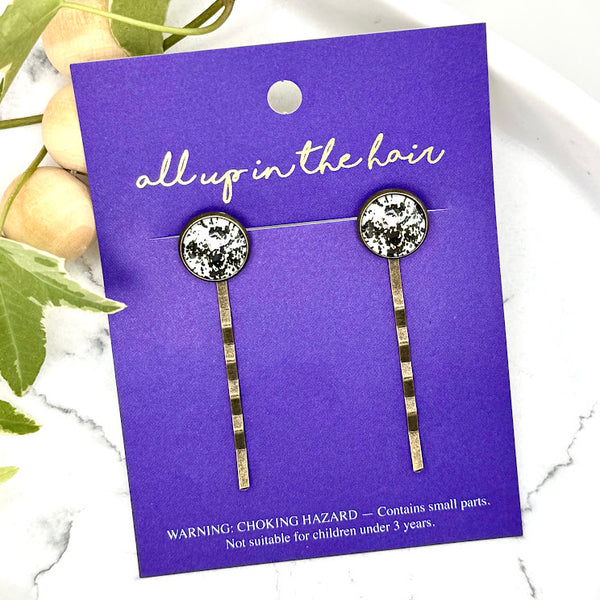 All Up In The Hair | Online Accessory Boutique Located in Mooresville, NC | Two cow print bobby pins on an All Up In The Hair branded packaging card. The card is laying on a gray background, surrounded by colorful glitter.