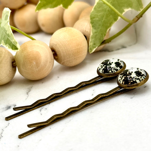 All Up In The Hair | Online Accessory Boutique Located in Mooresville, NC | Side view of two Cow Print Bobby Pins laying on a white marble background, next to a wood bead garland and ivy leaves.