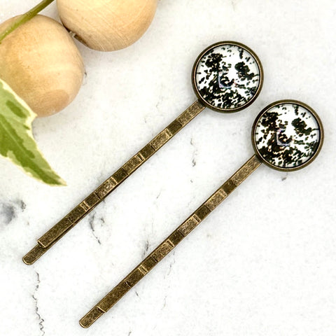 All Up In The Hair | Online Accessory Boutique Located in Mooresville, NC | Two Cow Print Bobby Pins laying diagonally on a white marble background, next to a wood bead garland and ivy leaves.