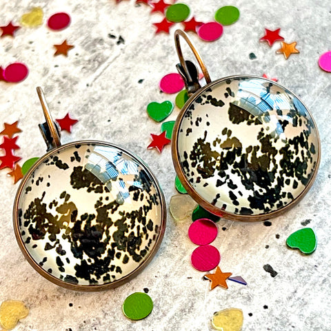 All Up In The Hair | Online Accessory Boutique Located in Mooresville, NC | Two cow print dangle earrings laying on a gray background, surrounded by colorful glitter.